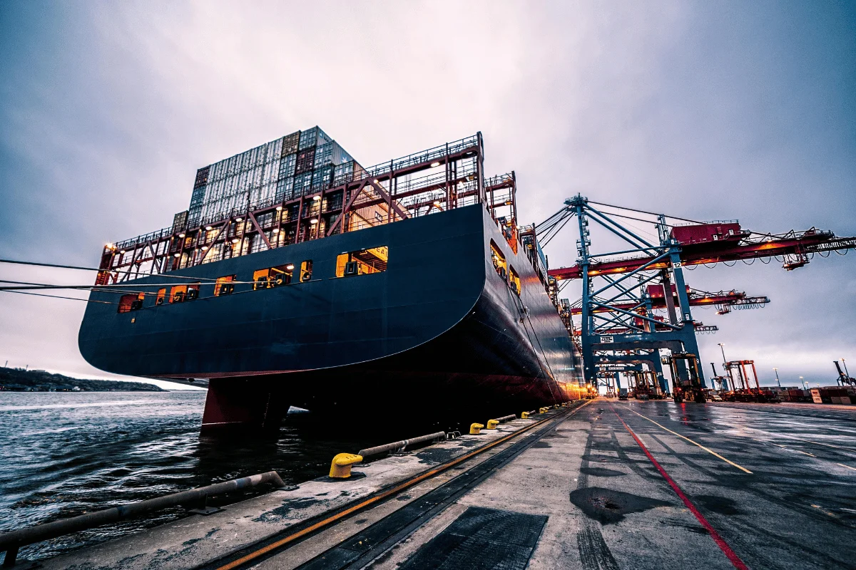A cargo ship docked at a port with containers being loaded by large cranes.