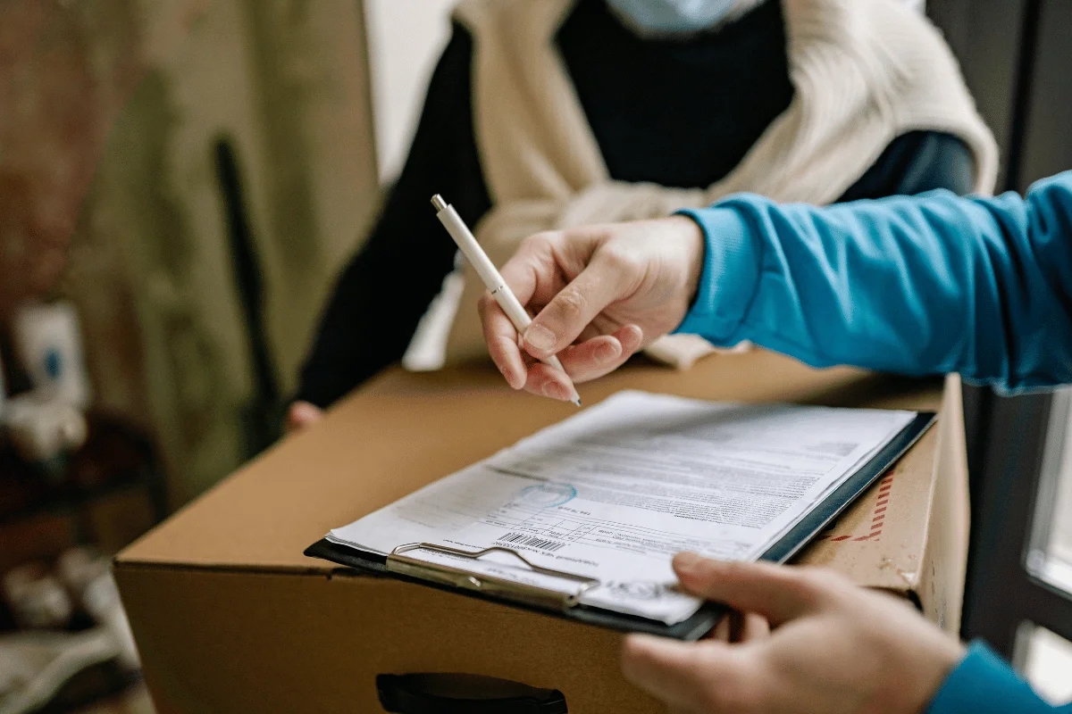 A person in a blue shirt signs a delivery note while holding a pen, next to a cardboard box, with another person in the background.