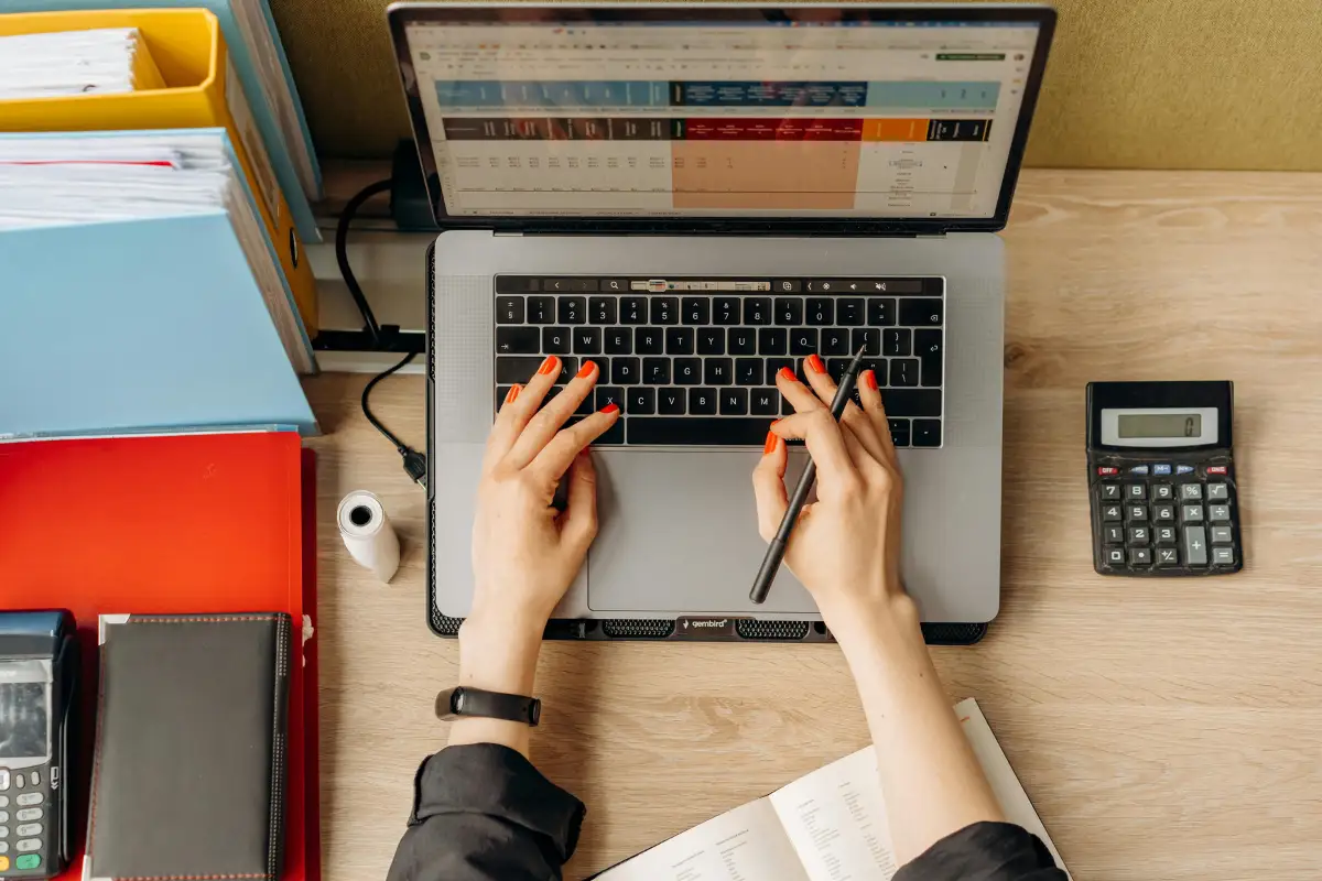 Desktop workspace with a laptop, calculator, colorful files, and a hand holding a pen.