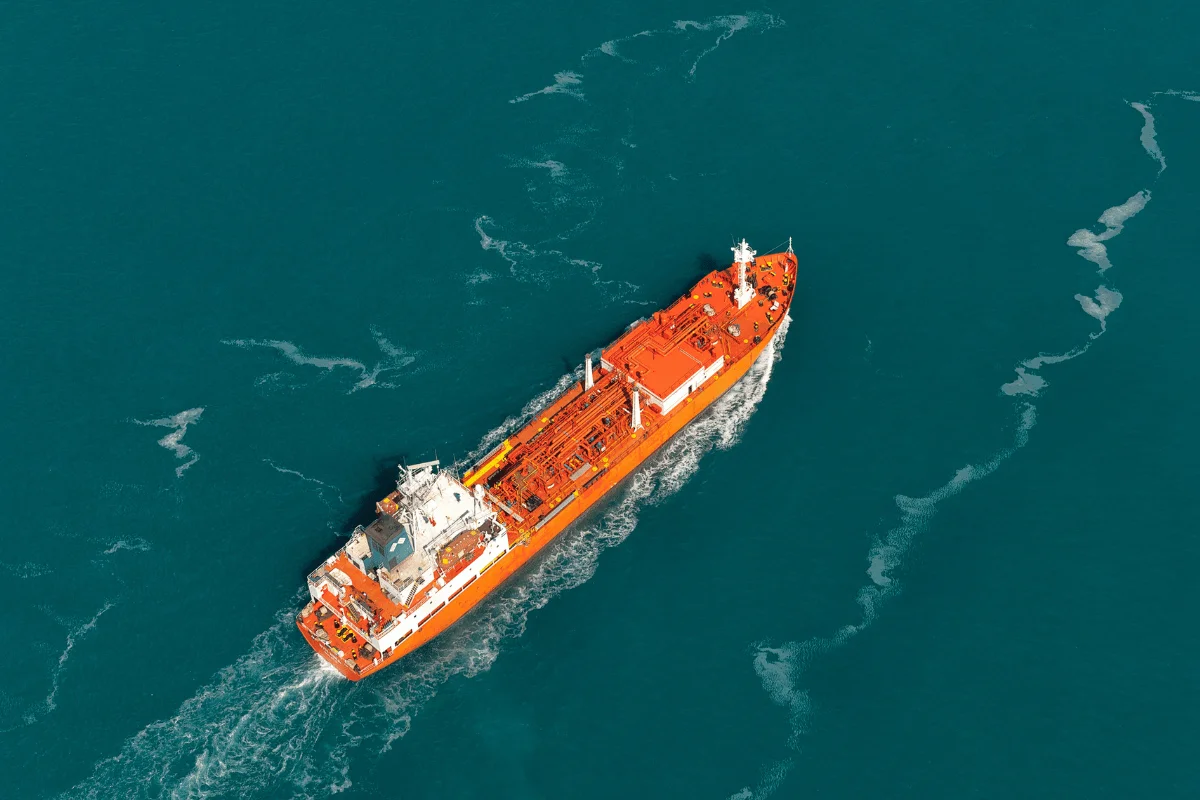 Aerial view of a large orange tanker ship navigating turquoise waters with a wake trail.