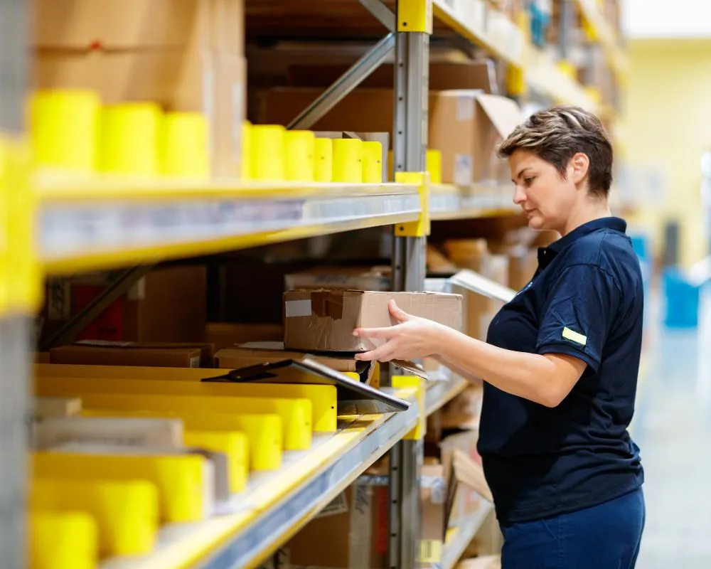 A warehouse worker organizing and inspecting packages on storage shelves. The scene represents inventory management, logistics operations, and supply chain efficiency in a fulfillment center.