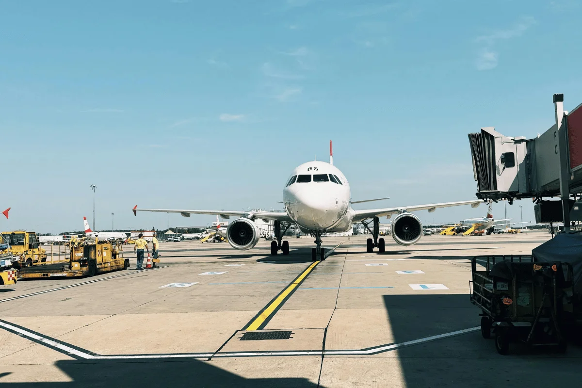 A white airplane with red tail markings taxis on a sunny airport runway, surrounded by ground equipment and other parked aircraft.