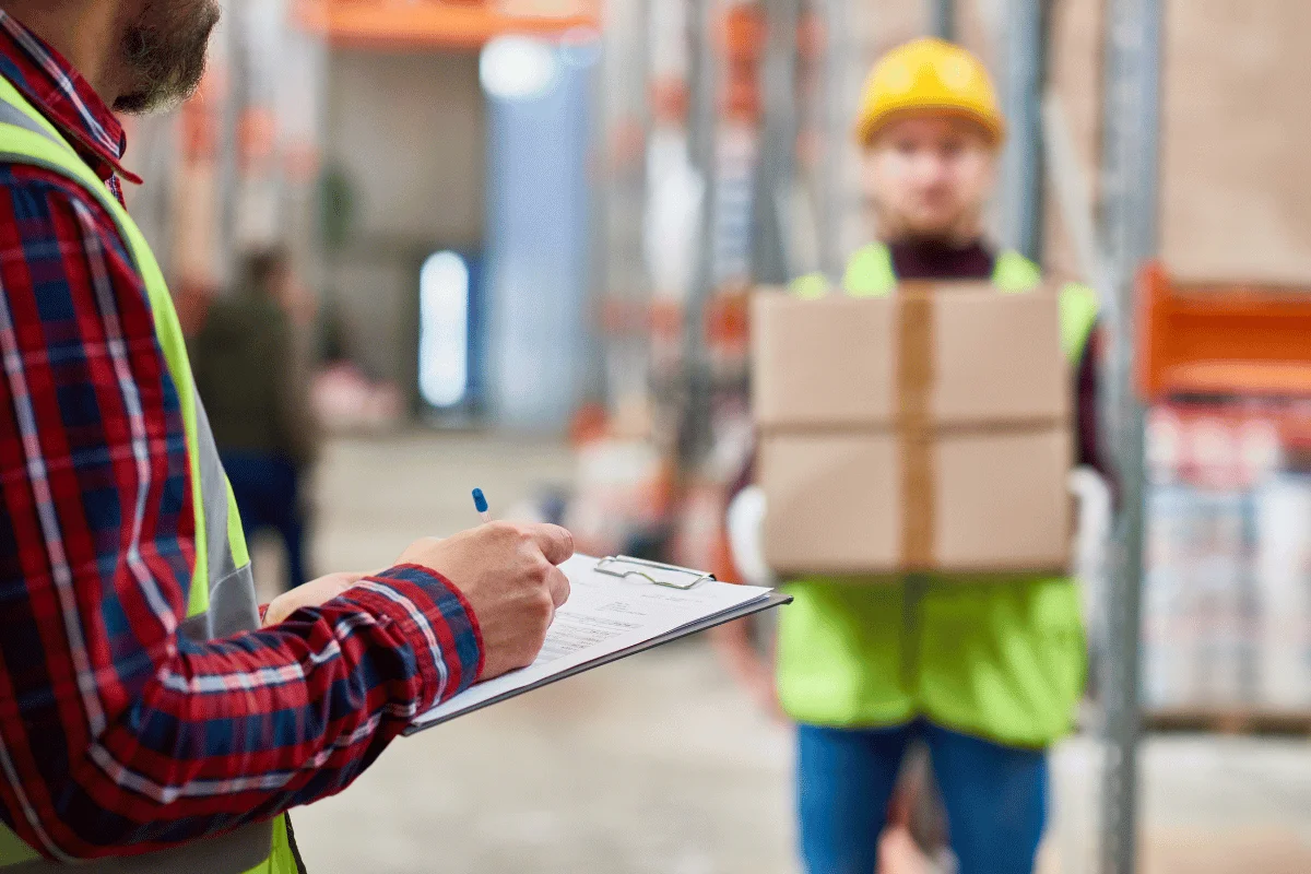 Logistics worker making notes on a clipboard with a delivery box being carried in the background.