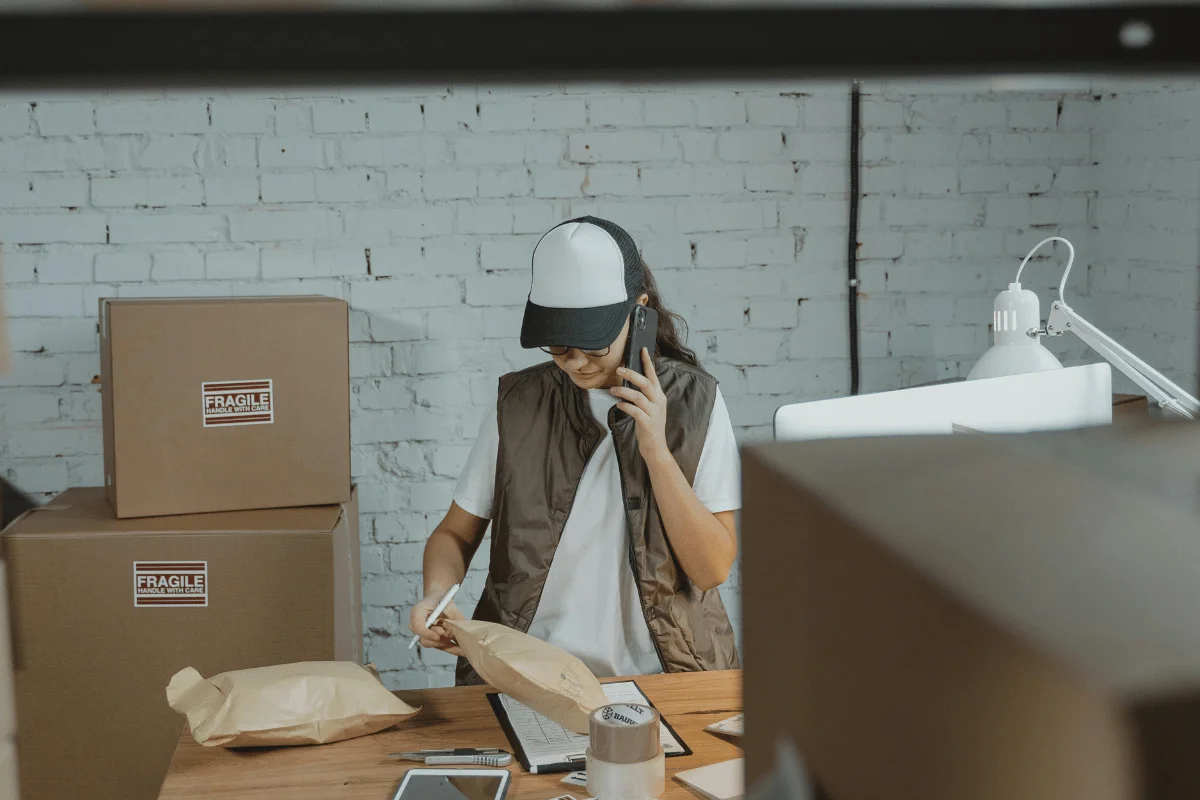 Woman wearing a cap and vest, handling packages and using a phone in a warehouse setting.