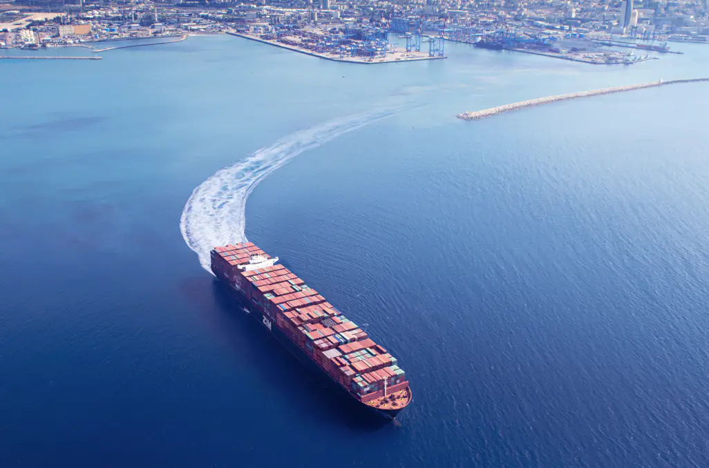 A large container ship making a turn in the open sea near a port, leaving a curved wake behind. The image represents global shipping, freight forwarding, and maritime logistics as the vessel navigates international trade routes.
