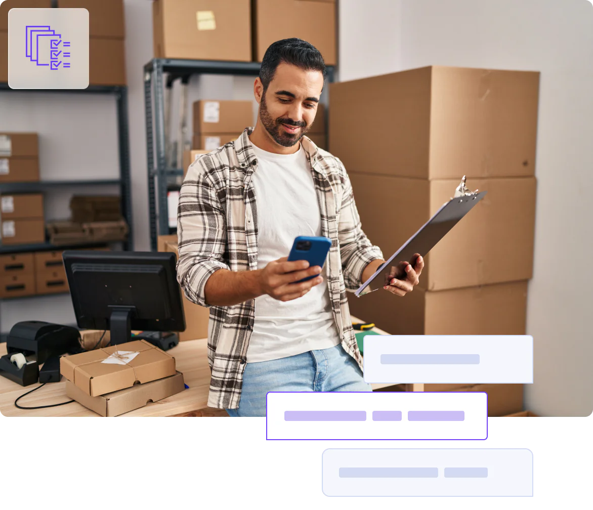 A person in a plaid shirt holding a clipboard and smartphone, with boxes and a computer in the background.