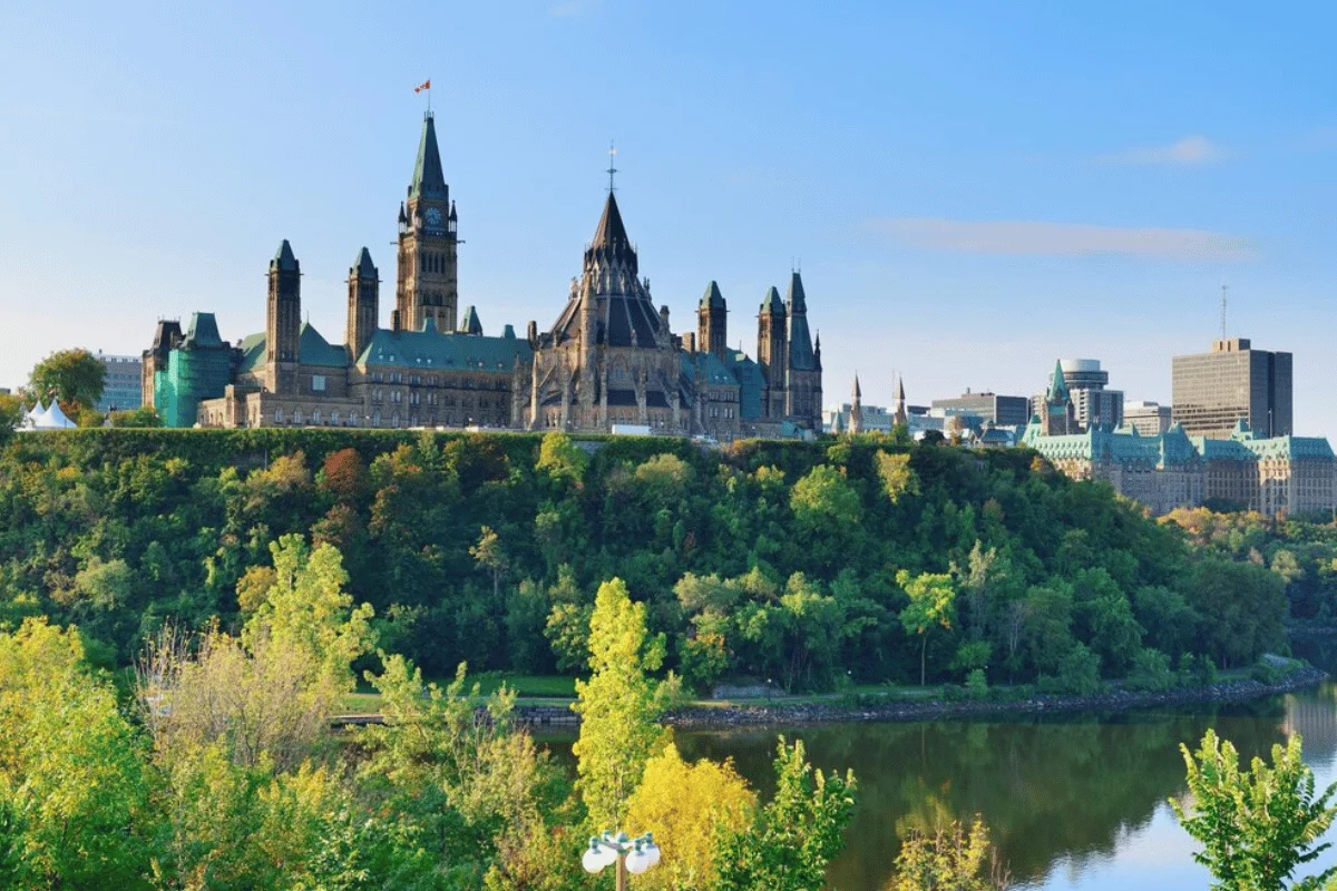 Ottawa's Parliament Buildings rise majestically above the Ottawa River, surrounded by lush greenery and a clear blue sky.
