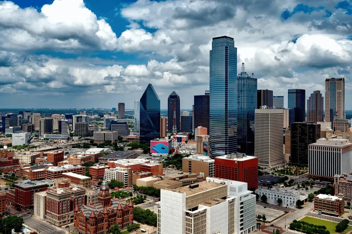 A panoramic view of the Dallas city skyline on a sunny day with scattered clouds.