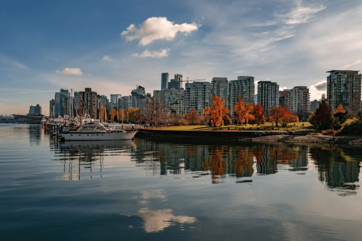 City skyline reflected in calm water, with boats docked and autumn trees in vibrant colors under a clear blue sky.