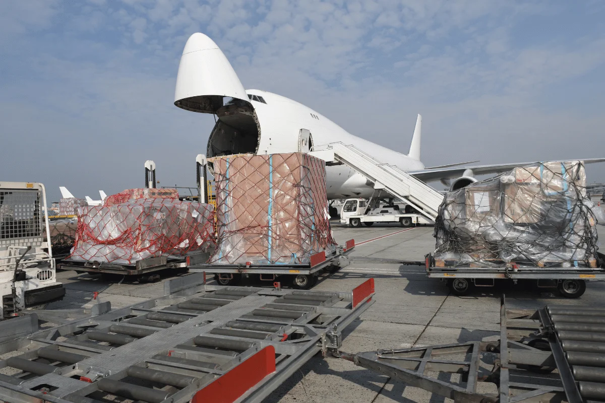 An aircraft with its nose open is being loaded with cargo wrapped in nets, while ground vehicles transport pallets nearby.