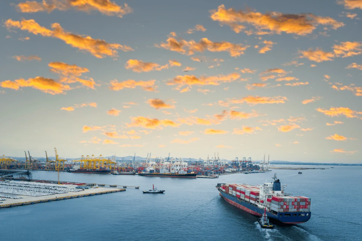 Cargo ship entering port at sunset with orange clouds and cranes in the background.