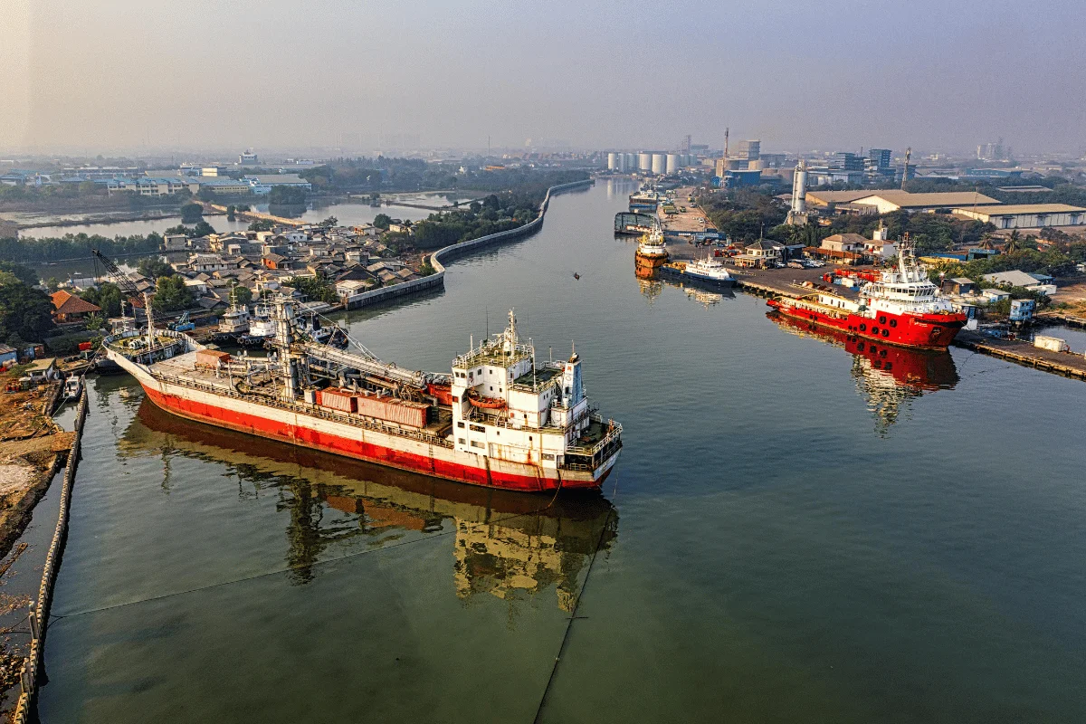 A busy shipping port in Indonesia with large vessels docked along the waterfront and surrounding industrial buildings.