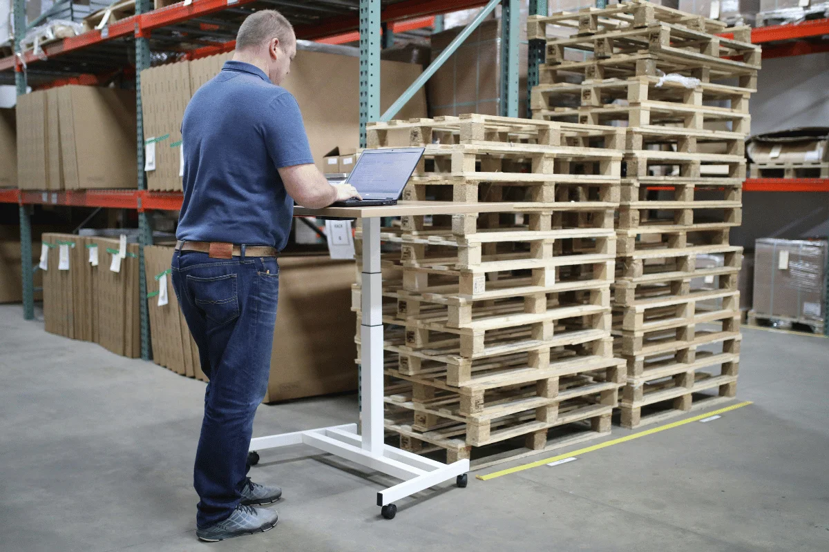 Warehouse manager using a laptop at a standing desk near stacked wooden pallets.