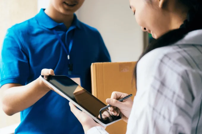 Woman using a stylus to sign for a package from a delivery person in a blue uniform.