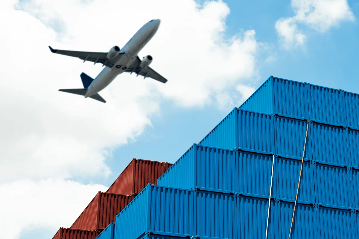 Freight Forwarders in Houston, TX: Services & Challenges 7 Airplane flying over stacked blue and red shipping containers against a cloudy sky.