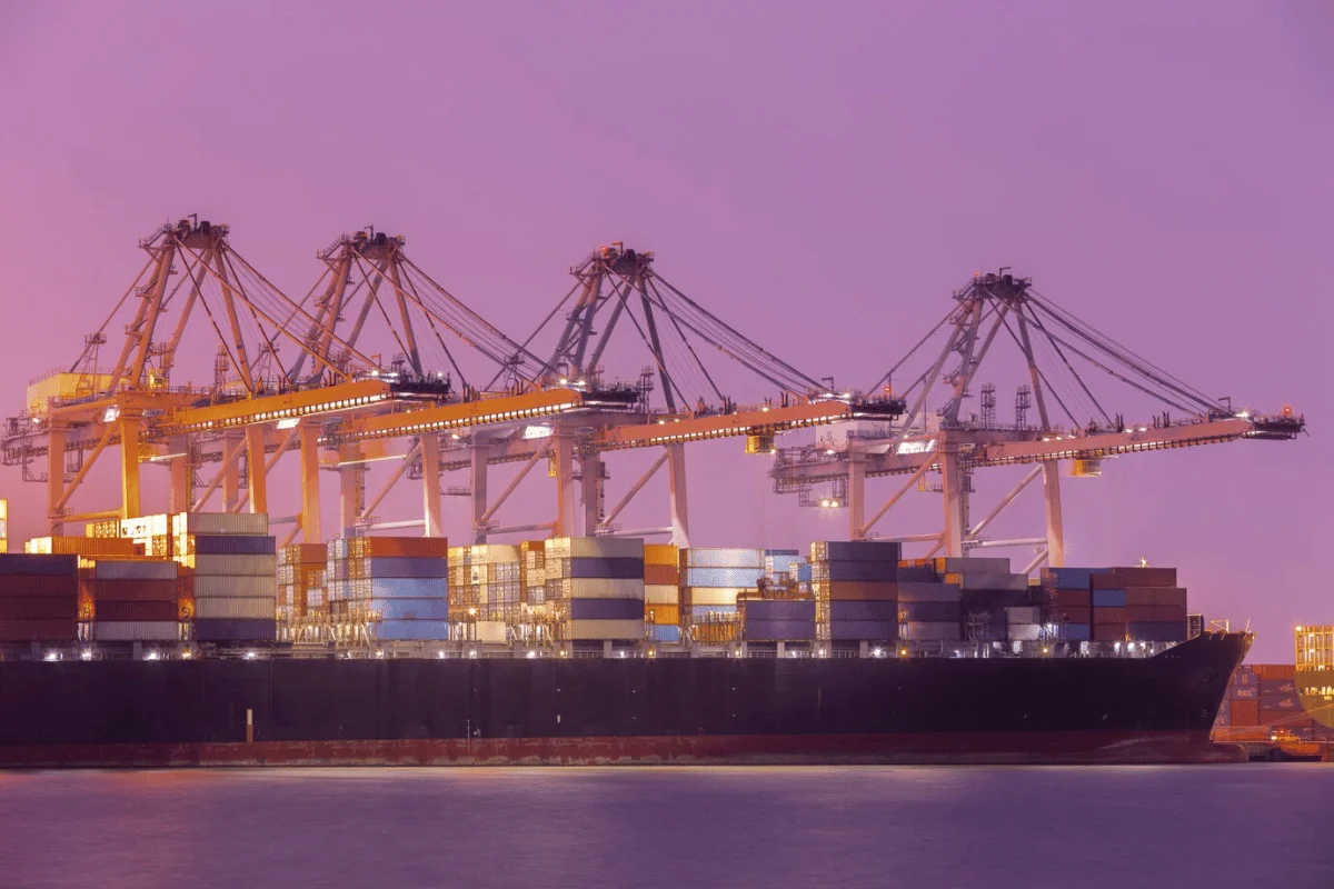 Container ship at a busy port with large cranes operating under a purple sky.