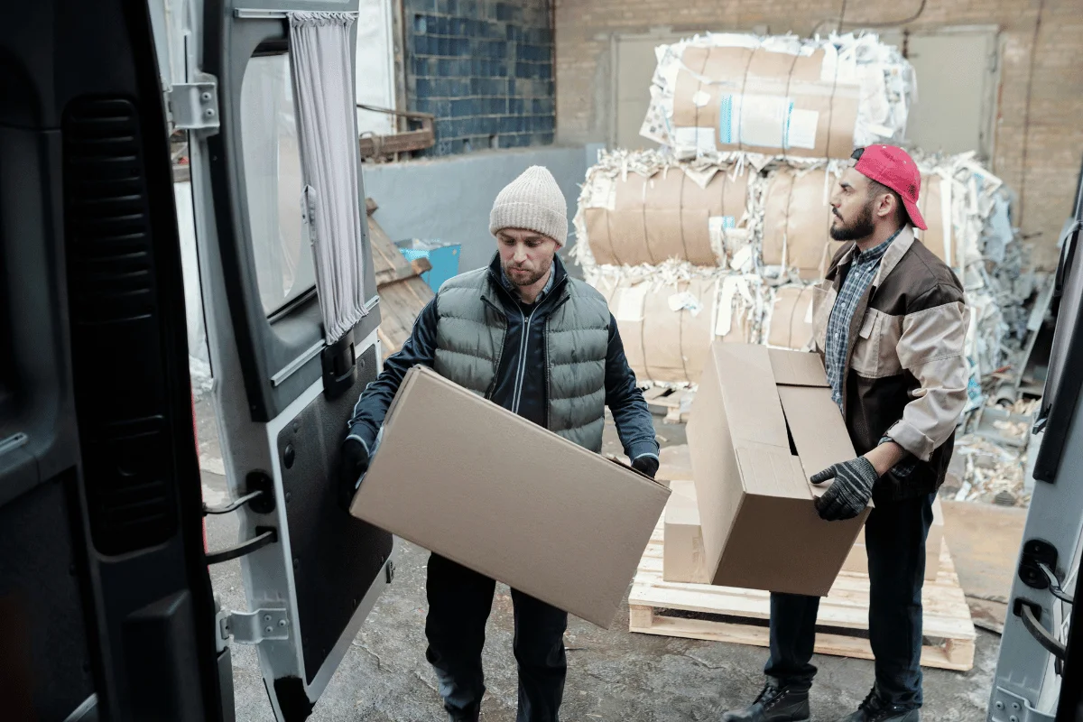 Freight Forwarder vs. Common Carrier: A Guide for Importers 11 Two workers unloading cardboard boxes from a delivery van at an industrial site with large paper rolls in the background.