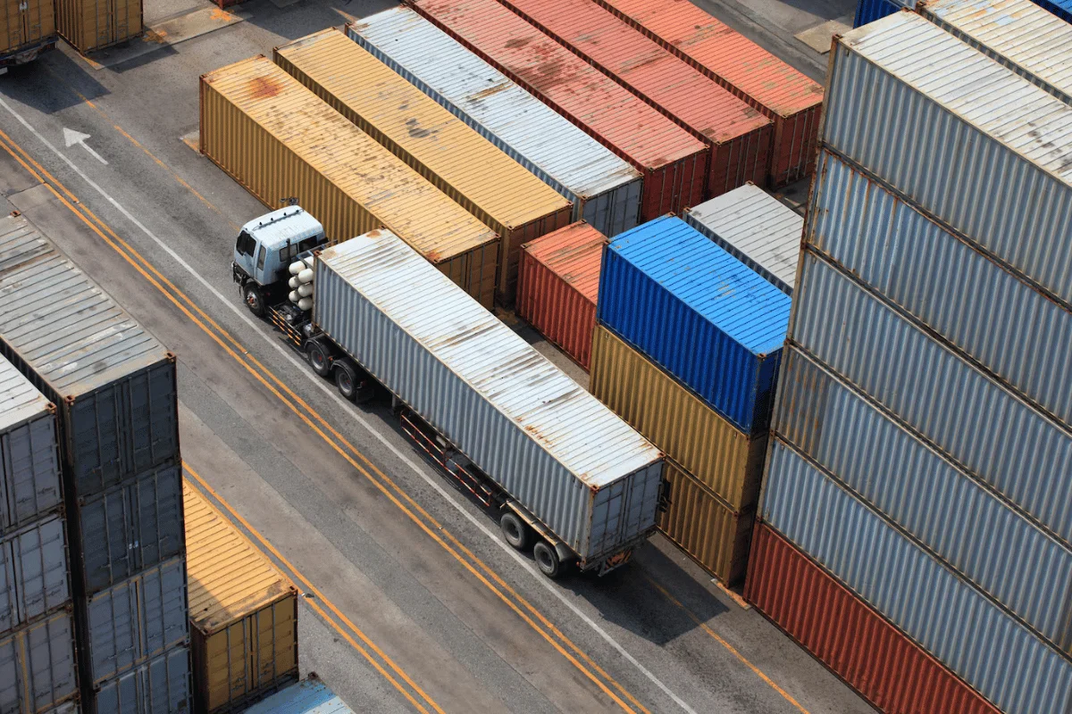 Truck driving through a shipping yard surrounded by stacked freight containers.